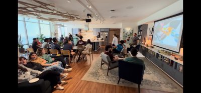 This photo shows Laurel cooking a chicken in a kitchen with a group of students watching and viewing it as well on a tv screen.